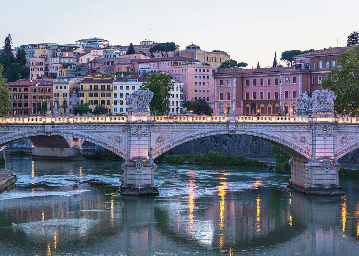A stone bridge over a calm river at dusk, with lights coming on along the bank
