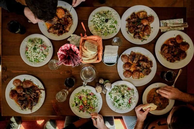 Plates of food on a long table seen from above, with hands reaching for dishes