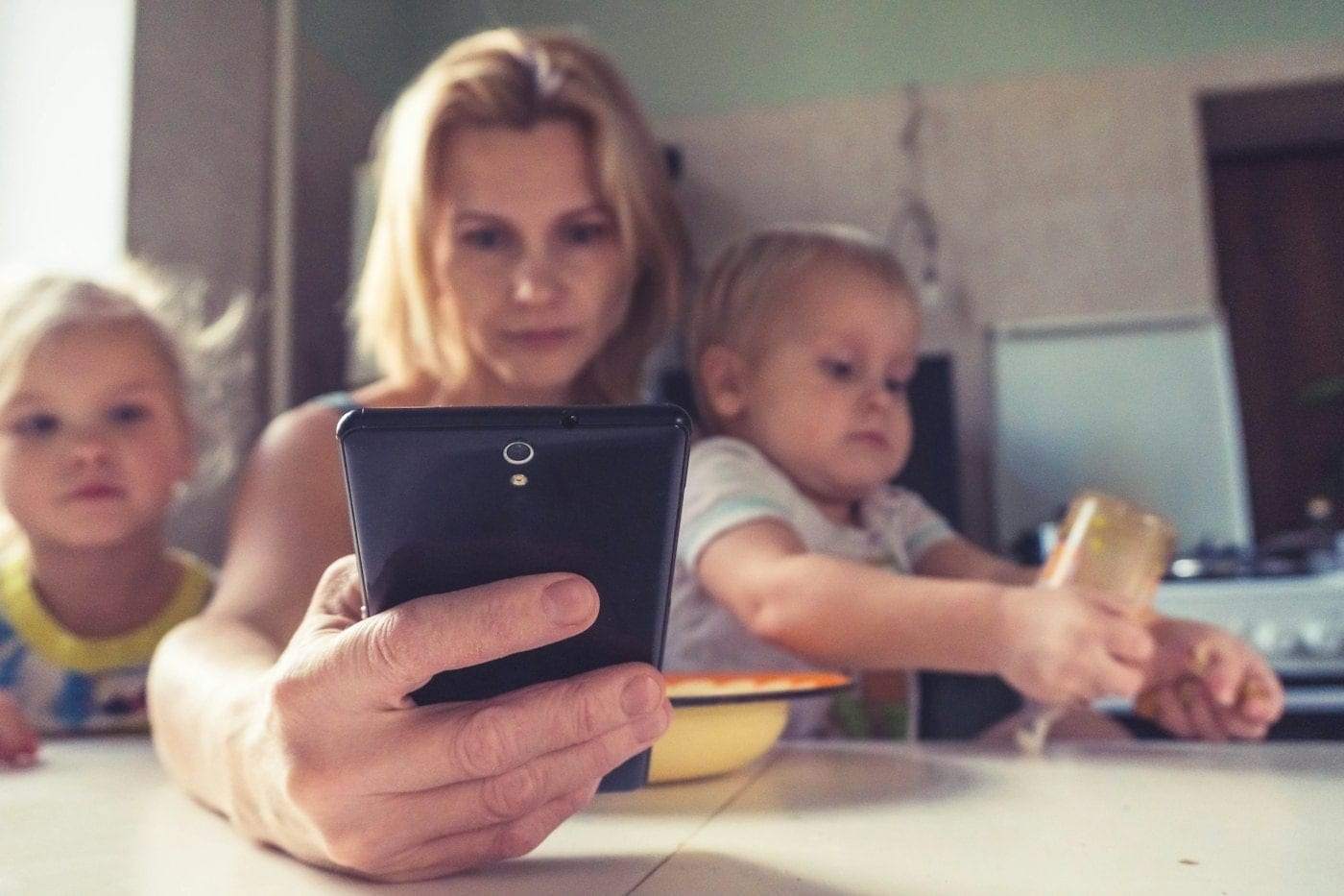 A mother holding up a tablet to take a photo while her two small children play beside her