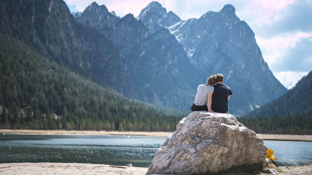 Two people sitting close together on a rock by a mountain lake